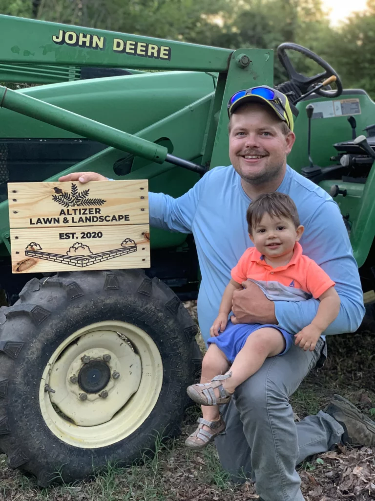 Man holding a wooden sign for Altizer Lawn & Landscape, established in 2020, while sitting with a young child beside a green John Deere tractor.